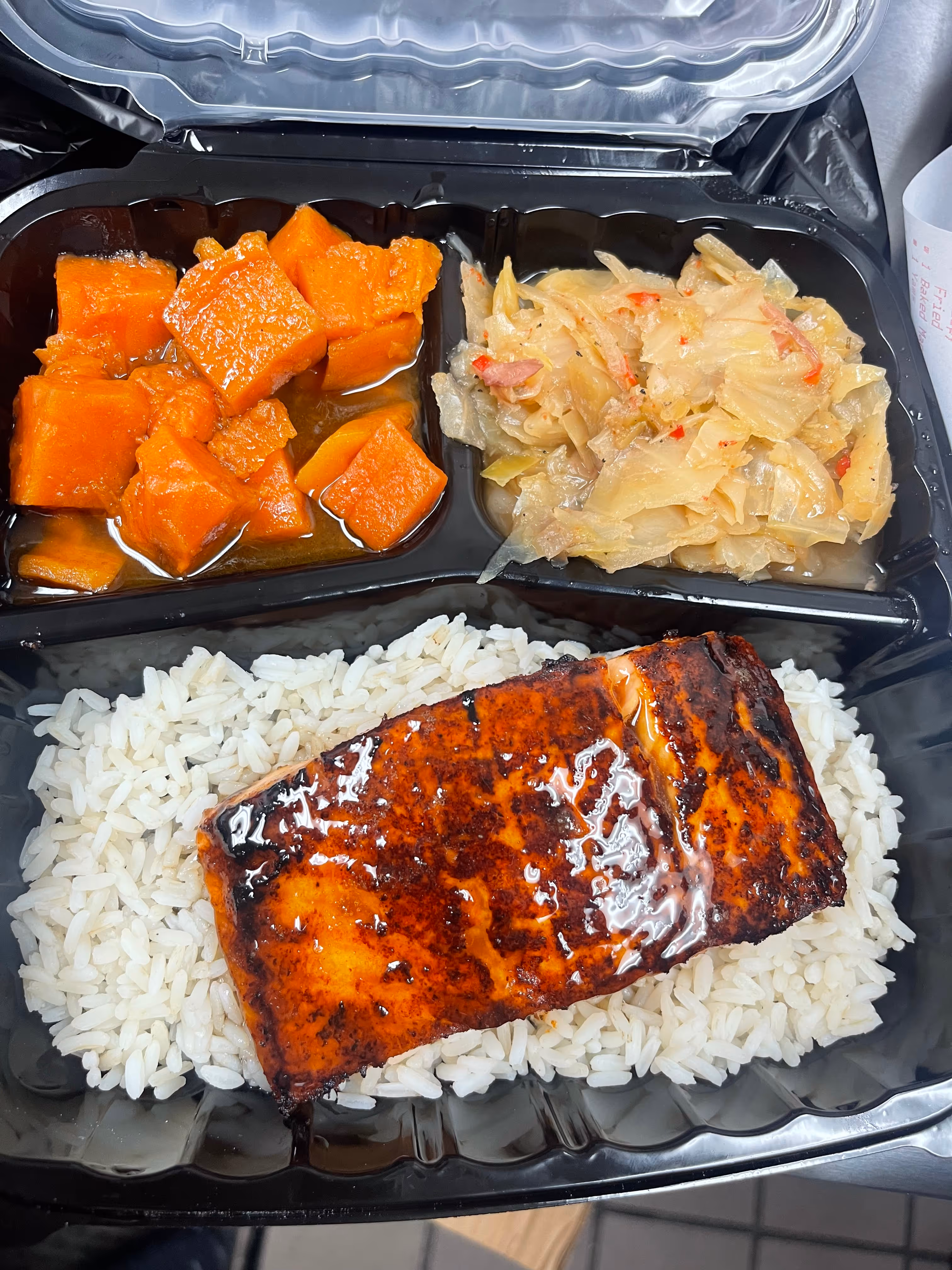 Tray with glazed grilled salmon on white rice, stewed sweet potatoes, and cooked cabbage with peppers.