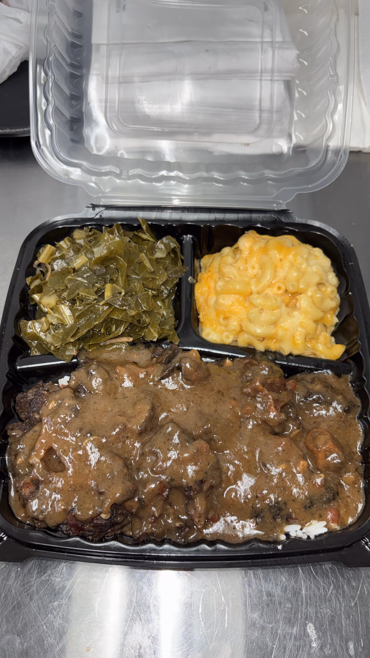 Takeout container with smothered meat in brown gravy over rice, cooked greens, and macaroni and cheese.