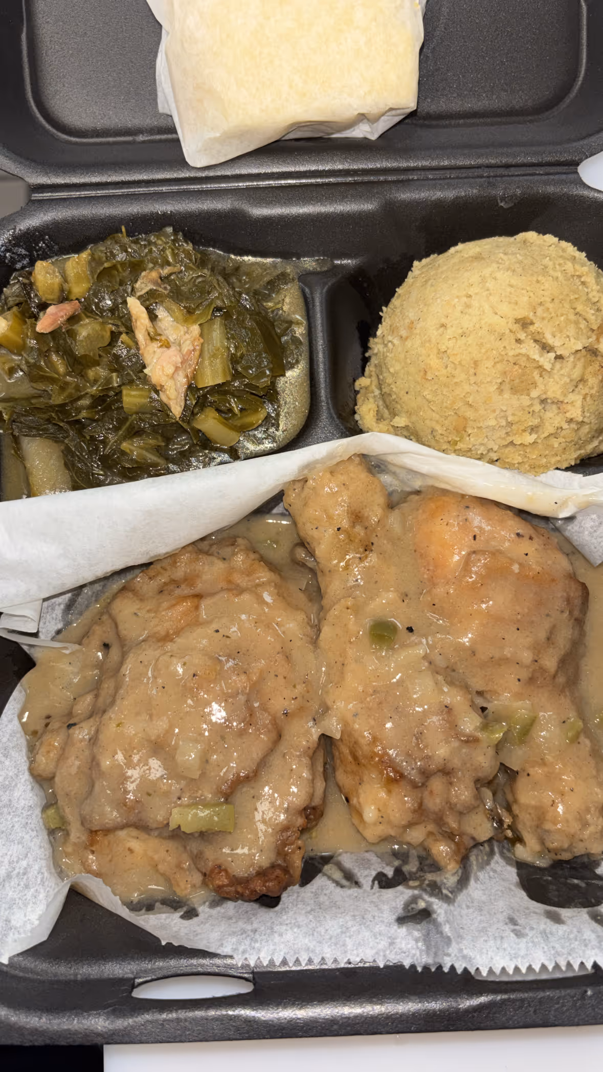 Takeout meal with two fried chicken pieces covered in gravy, a scoop of mashed potatoes, and cooked greens in a black plastic container.