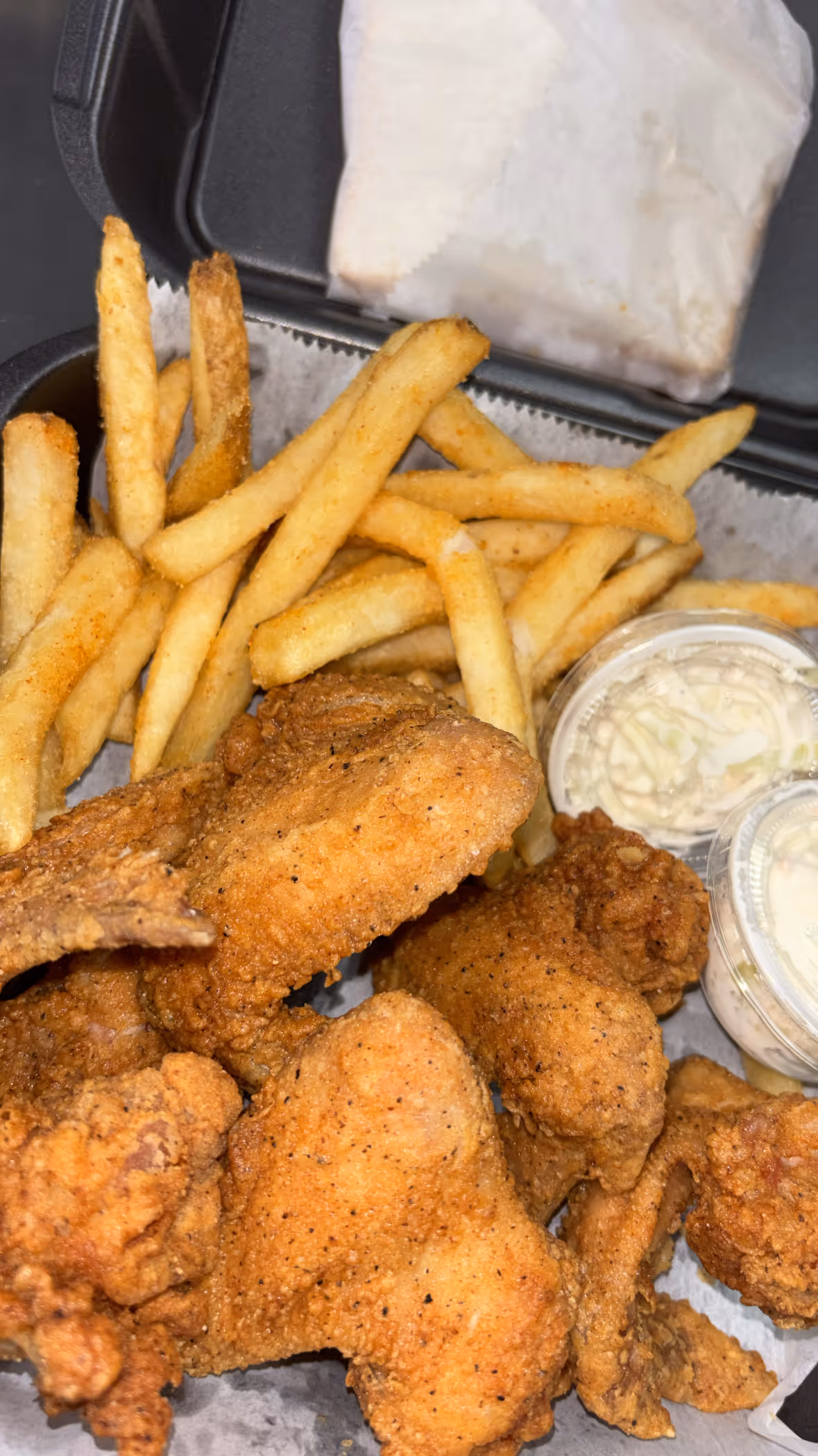 Takeout box filled with seasoned French fries, fried chicken wings, two containers of coleslaw, and a wrapped sandwich.