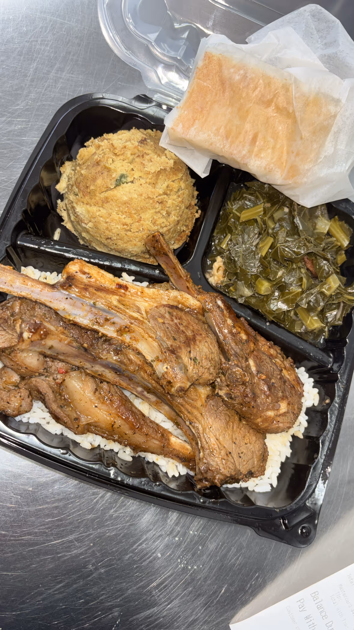 Takeout meal with grilled lamb chops on white rice, cornbread muffin, collard greens, and a wrapped biscuit in a black plastic container.