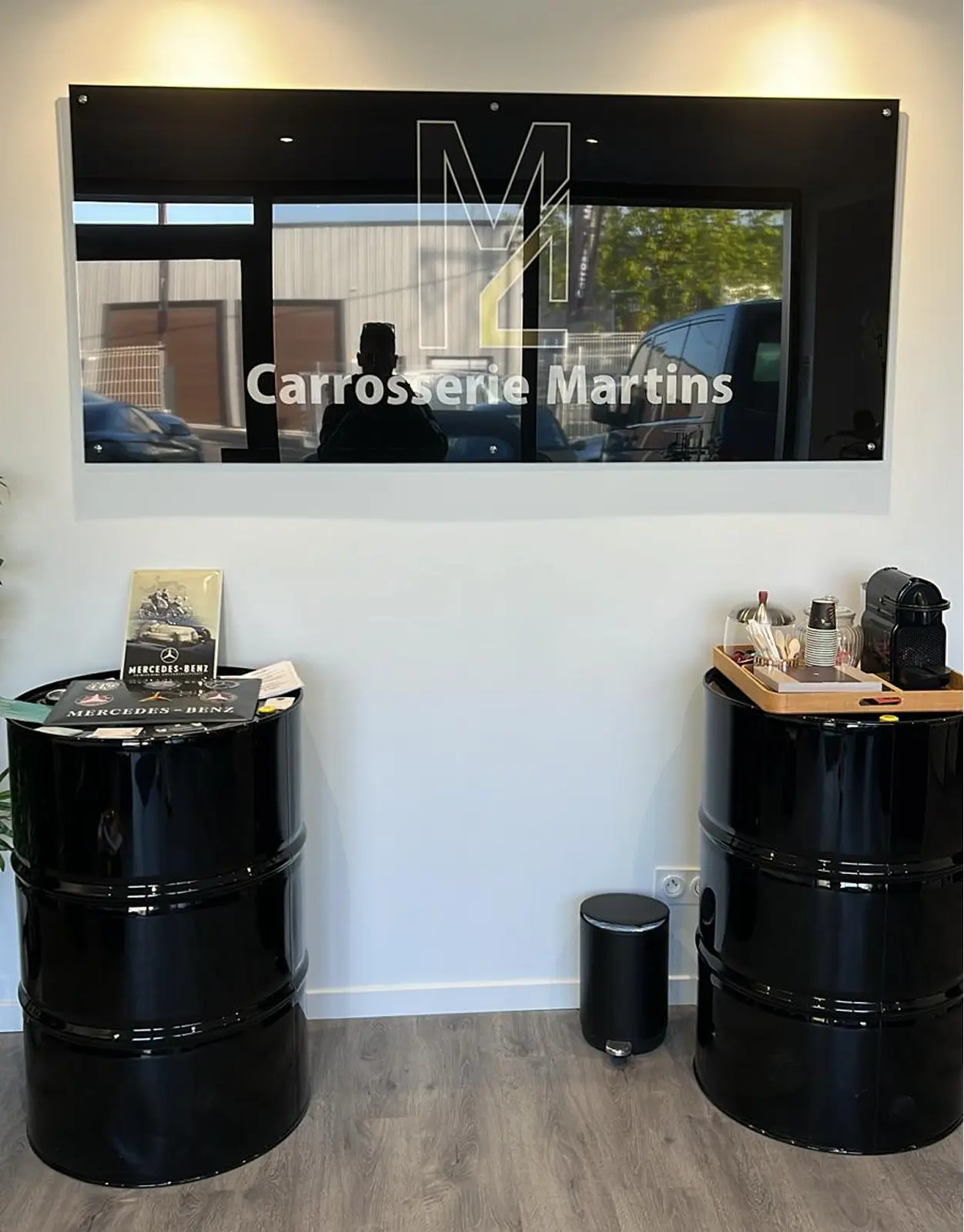 Interior of Carrosserie Martins shop with a black sign on the wall, two black barrel tables, one with Mercedes-Benz brochures and the other with a coffee machine and cups.
