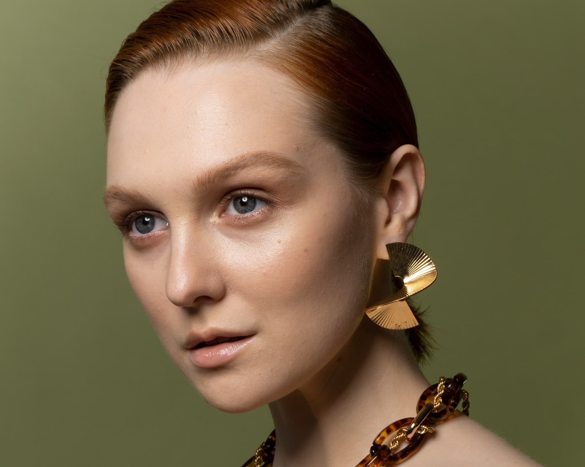 Close-up of a woman with slicked-back auburn hair wearing large gold geometric earrings and a chunky tortoiseshell necklace, against an olive green background.