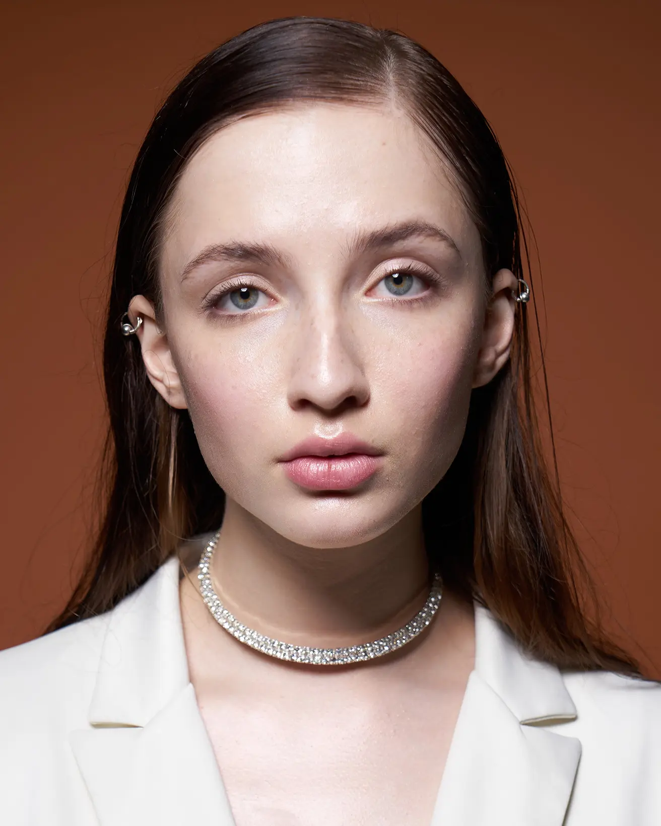 Close-up portrait of a woman with light blue eyes, wearing a white blazer and a sparkling diamond choker necklace, against a brown background.