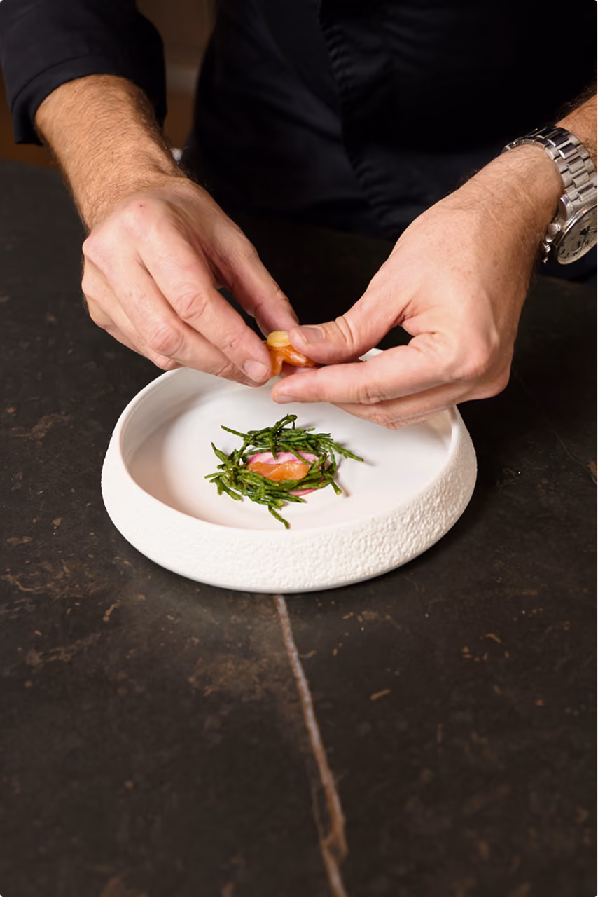 Chef Gábor Viczián's hands plating fish with samphire in a white textured bowl.