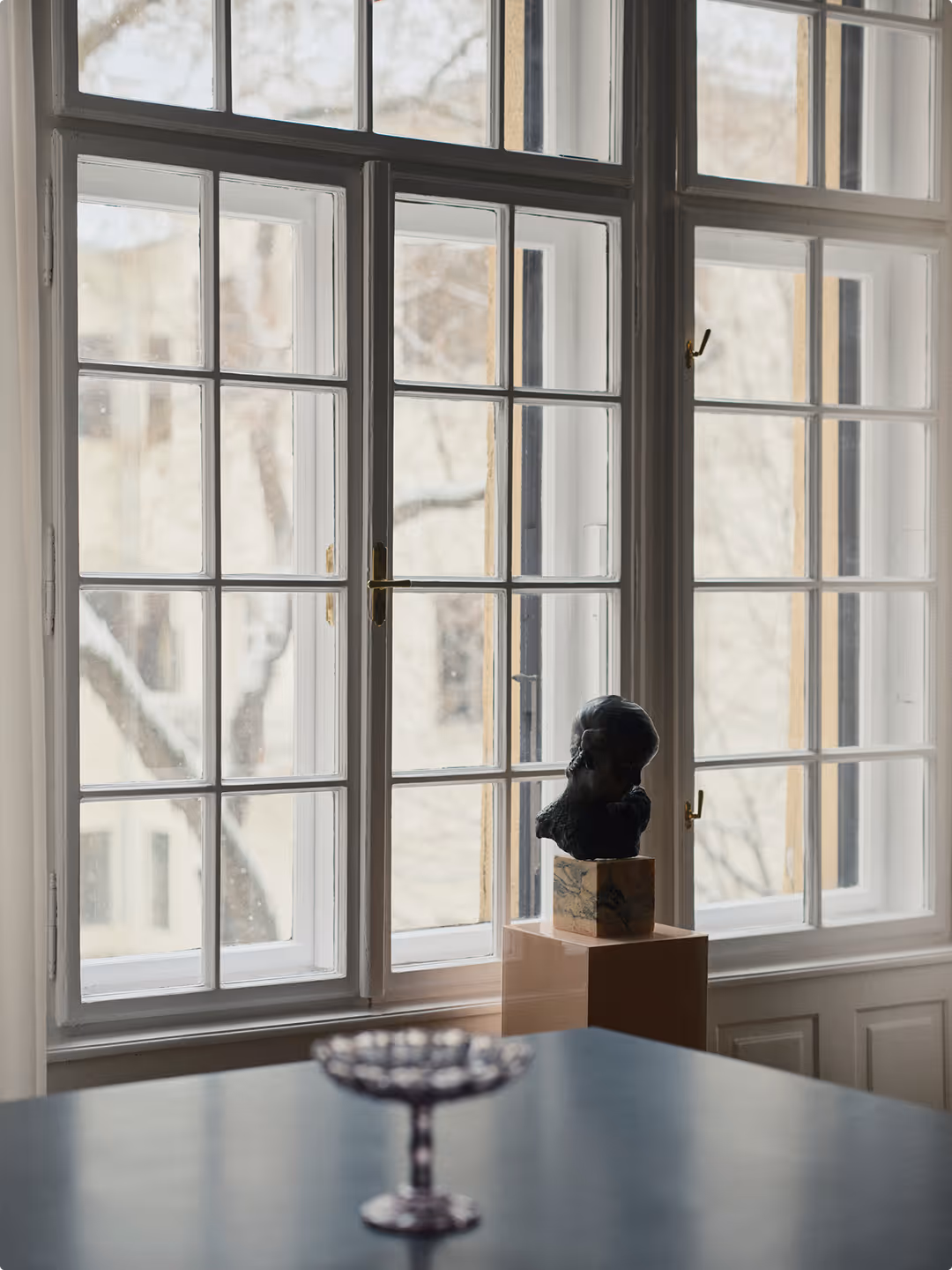 A dark bust on a plinth in front of a tall paned window, with a silver footed bowl on a dark table in the foreground.