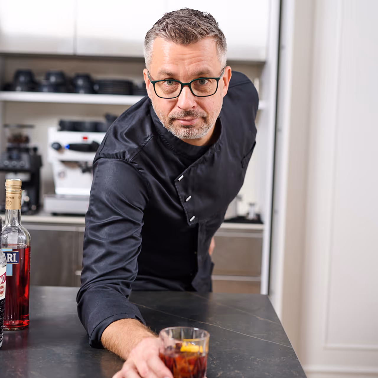 Chef Gábor Viczián in a black chef's jacket leaning on the counter beside a cocktail garnished with orange.