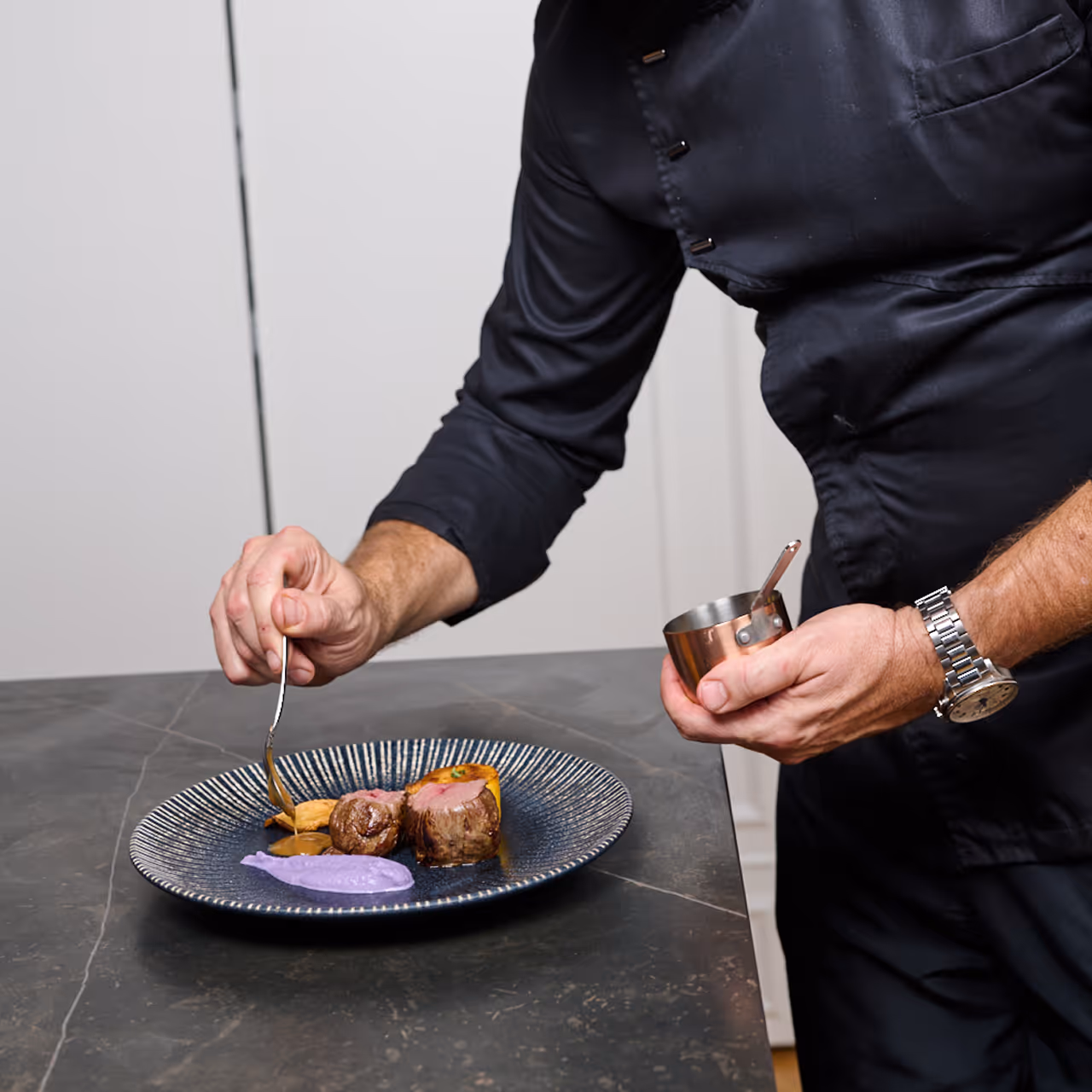 Chef Gábor Viczián spooning sauce over a meat course with purple purée on a dark ribbed plate.