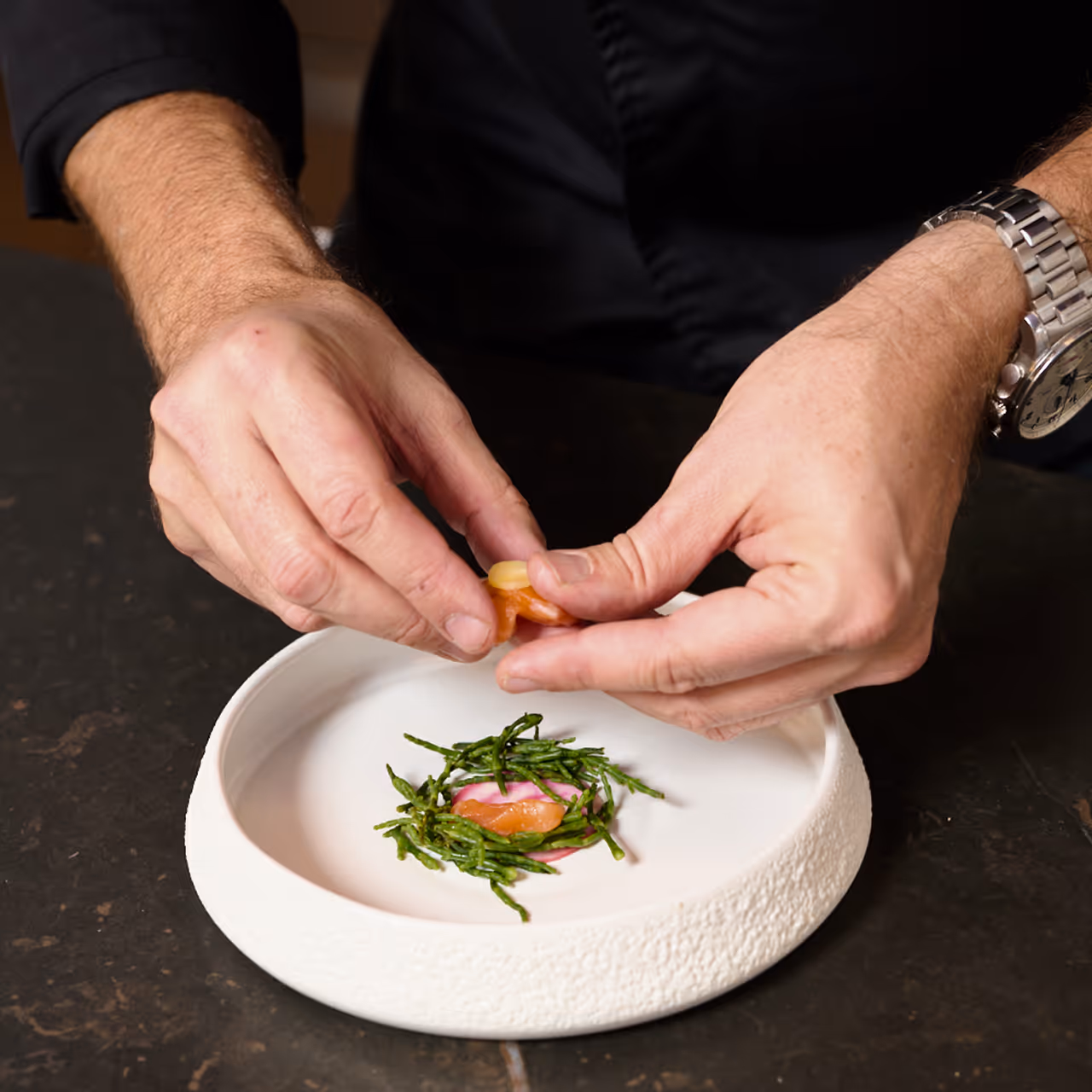 Chef Gábor Viczián's hands plating fish with samphire in a white textured bowl.