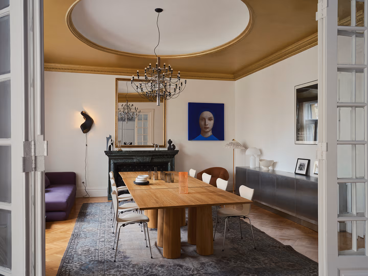 The dining room seen through open double doors: a long communal wooden table with white chairs, a chandelier, a black fireplace and a portrait on the wall.