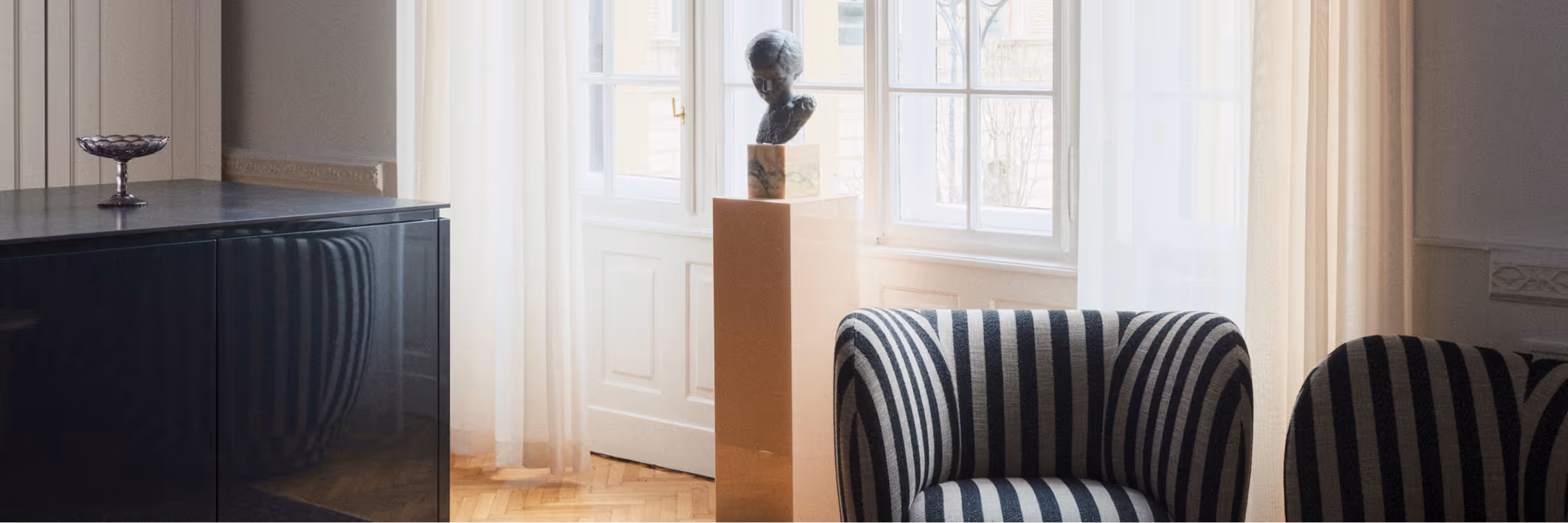 A bright room with tall windows, a dark sideboard, a bust on a plinth and two black-and-white striped armchairs.