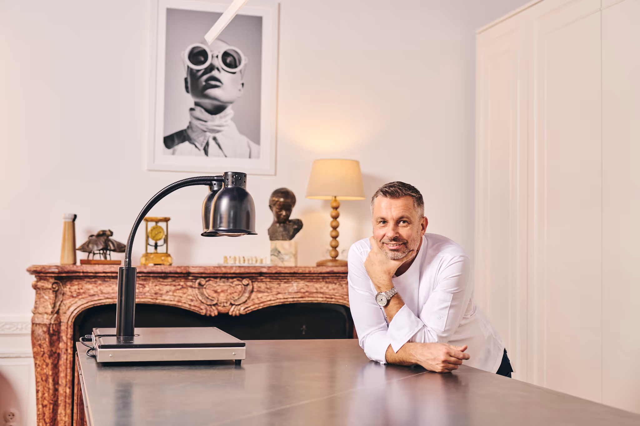 Chef Gábor Viczián in a white chef's jacket leaning on the kitchen counter, with a red marble fireplace and a black-and-white fashion photo behind him.
