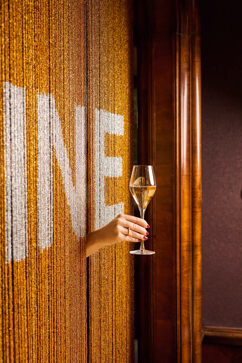 A hand holding a glass of champagne out through a gold beaded curtain spelling “SHINE”.