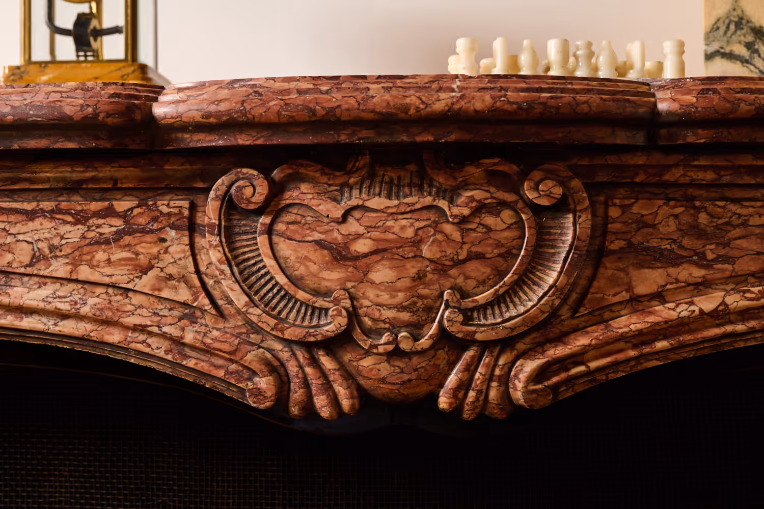 Close-up of a carved red marble mantelpiece with a gold clock and white chess pieces on top.