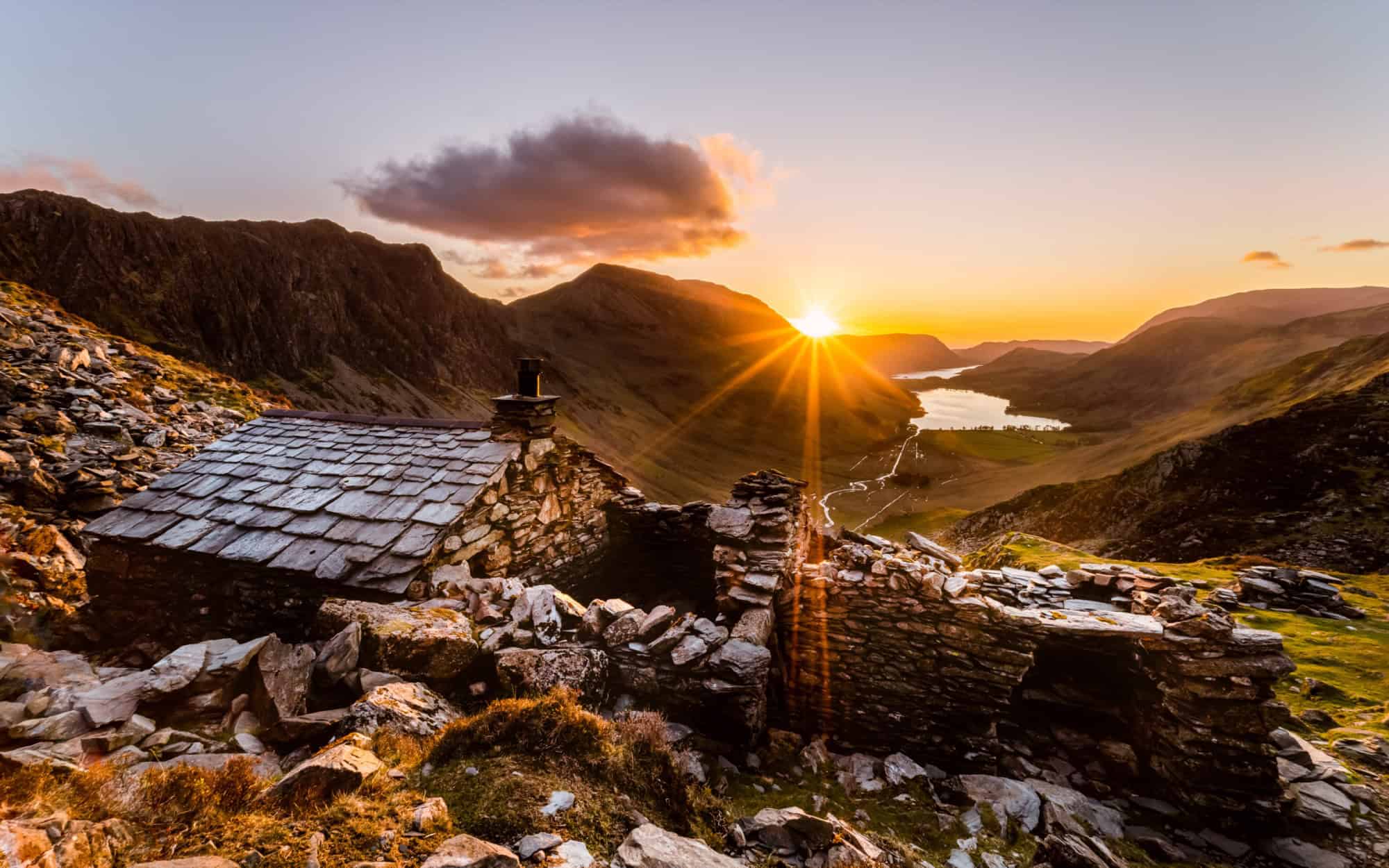 Sunset over the wild Cumbrian landscape with a historic stone bothy and rolling green valleys.
