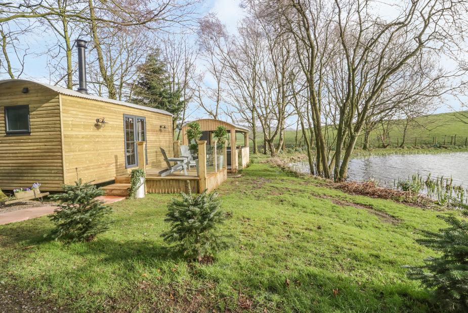 High Rigg Shepherd's Delight wooden lodge beside a lake with deck chairs and trees nearby