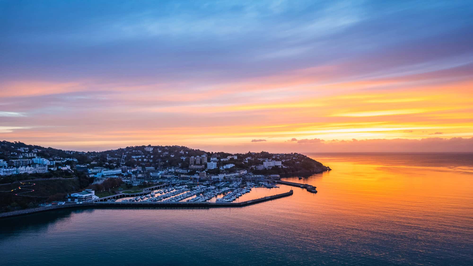 Aerial view of Torquay Marina at sunrise, with golden light reflecting off the calm sea and luxury yachts moored in the harbour.