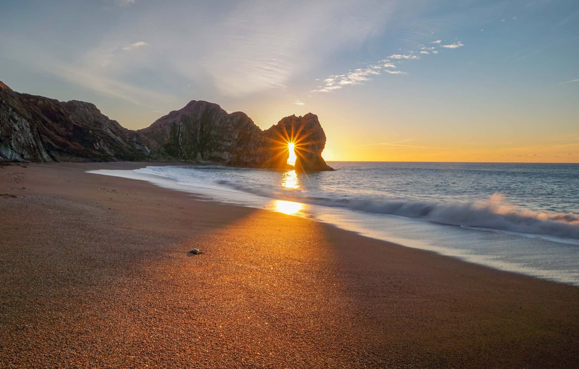 Golden sunrise at Durdle Door in Dorset, featuring a dramatic sunburst shining through the iconic limestone arch onto the sandy beach.