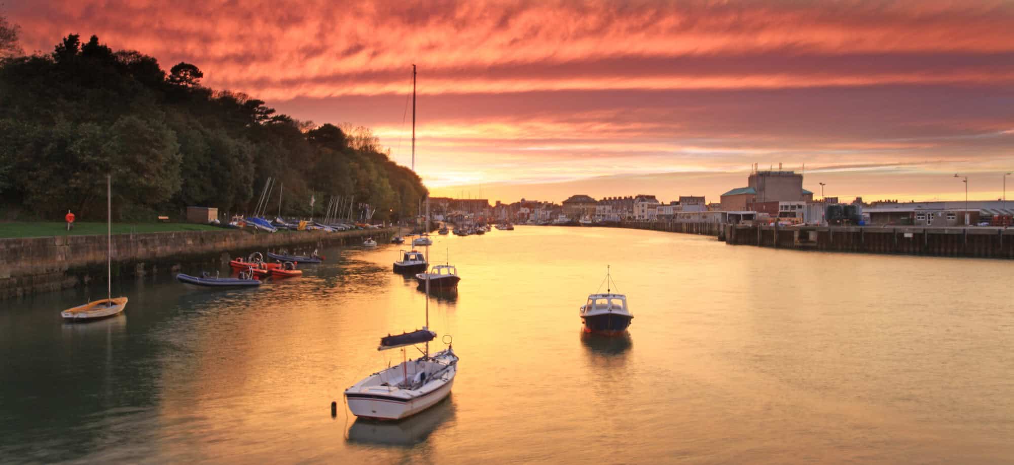 Sunset over the historic Weymouth Harbour, featuring moored boats and the townscape bathed in warm evening light.