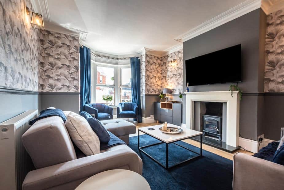 Cottage living room with fireplace, wall-mounted TV, and bay window with blue chairs