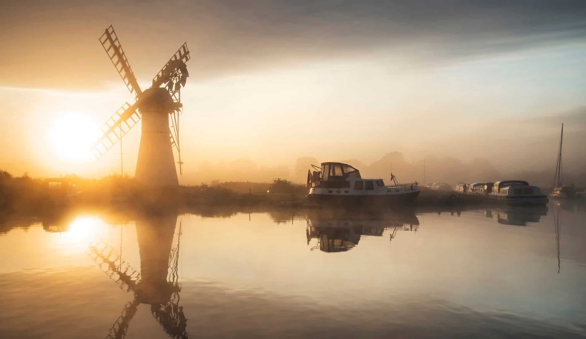 Golden sunrise mist rising over the calm river and Thurne Mill windmill in the Norfolk Broads, East of England.