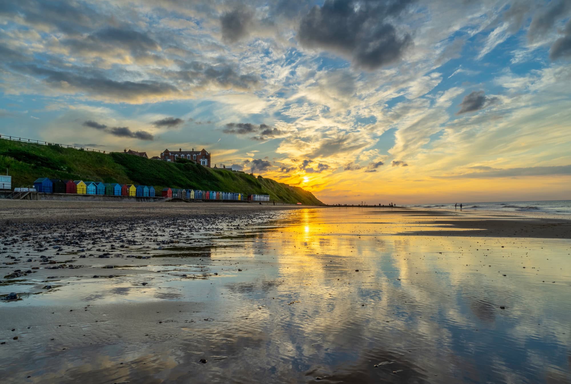 Golden sunset reflecting on the vast wet sands of Mundesley Beach in Norfolk, with colourful beach huts on the cliffs.