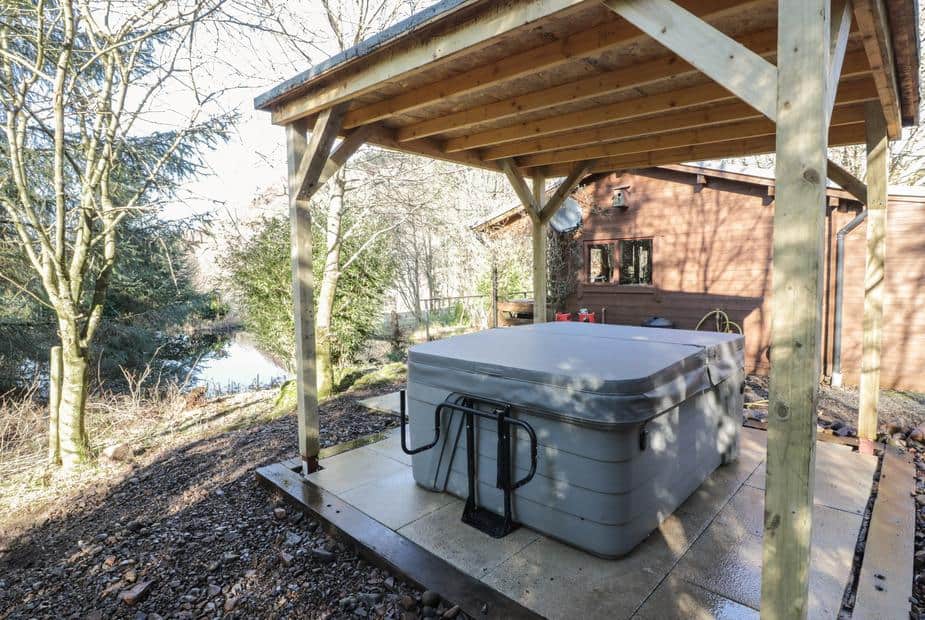 Covered hot tub beside lodge with trees and a small lake in the background.