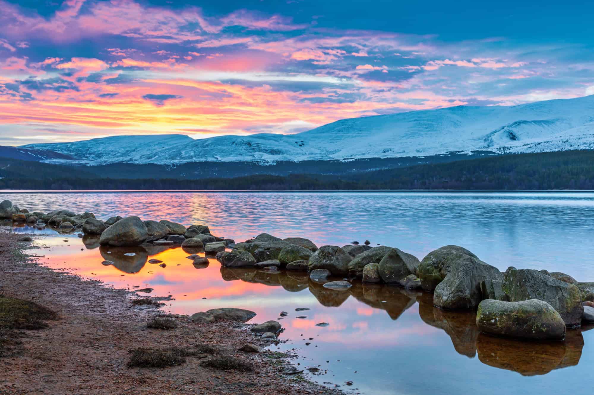 Vibrant pink sunrise reflecting in Loch Morlich with snow-capped Cairngorm mountains in the background.