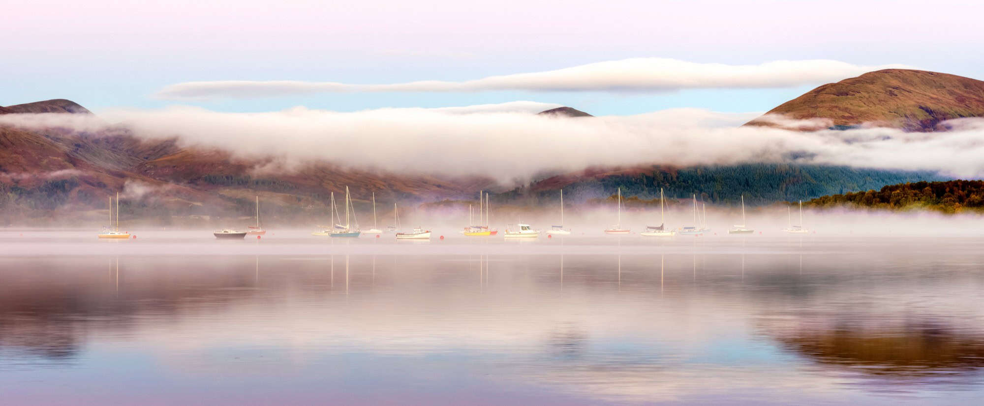 Pastel sunrise with mist rising over the calm waters and moored boats at Milarrochy Bay, Loch Lomond.