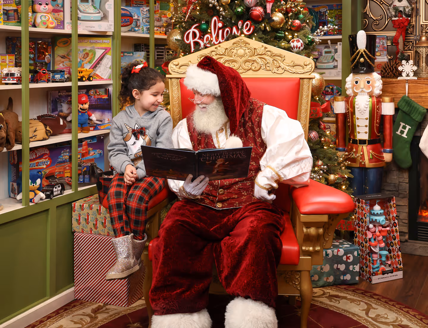 Child reading Christmas book with Santa on golden throne in toy-filled holiday workshop