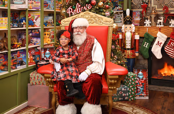 Two children sitting with Santa on golden throne in festive workshop with toy shelves and fireplace