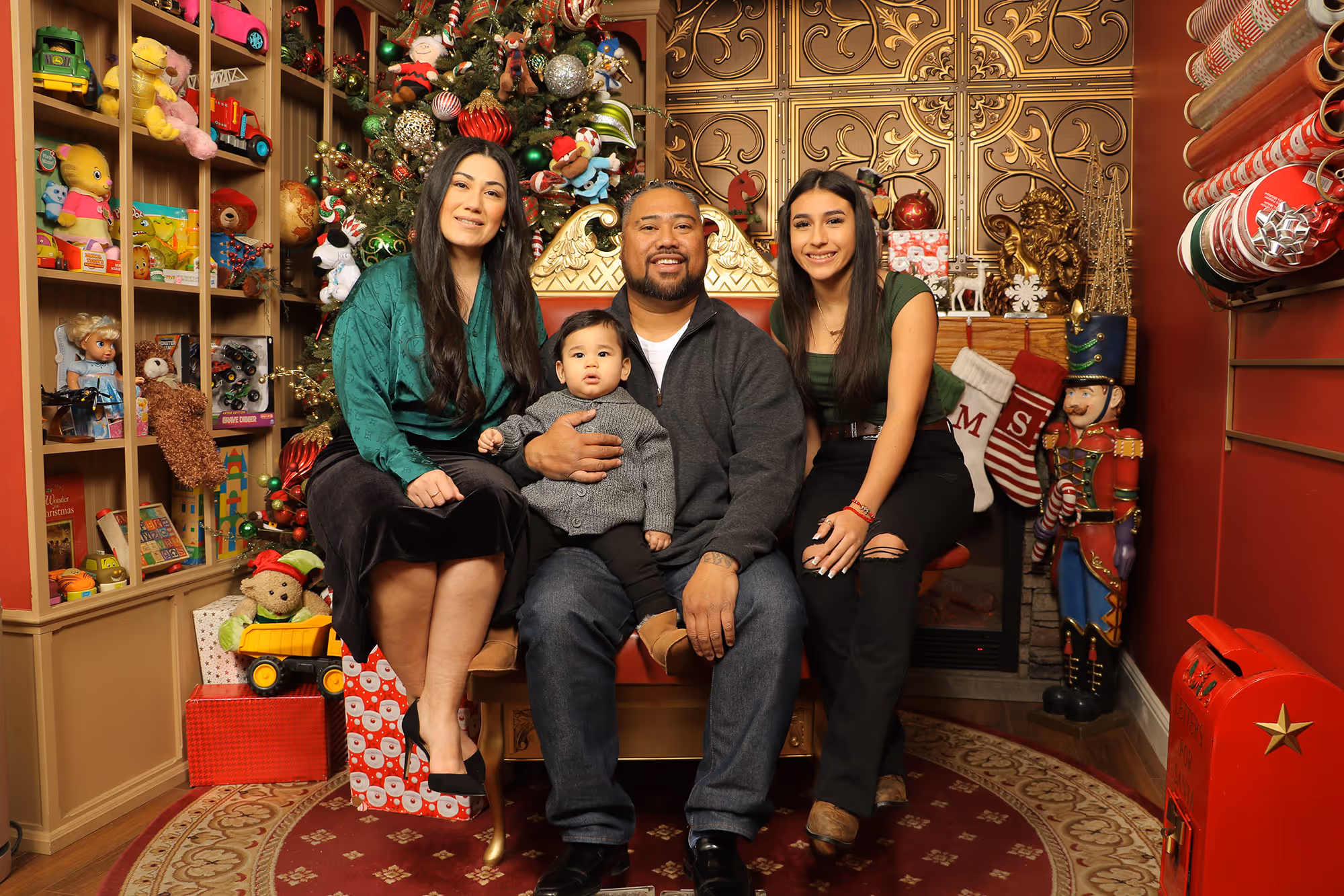 Family of four pictured in a Christmas photo studio that is festively-decorated with presents, ornaments and nutcrackers