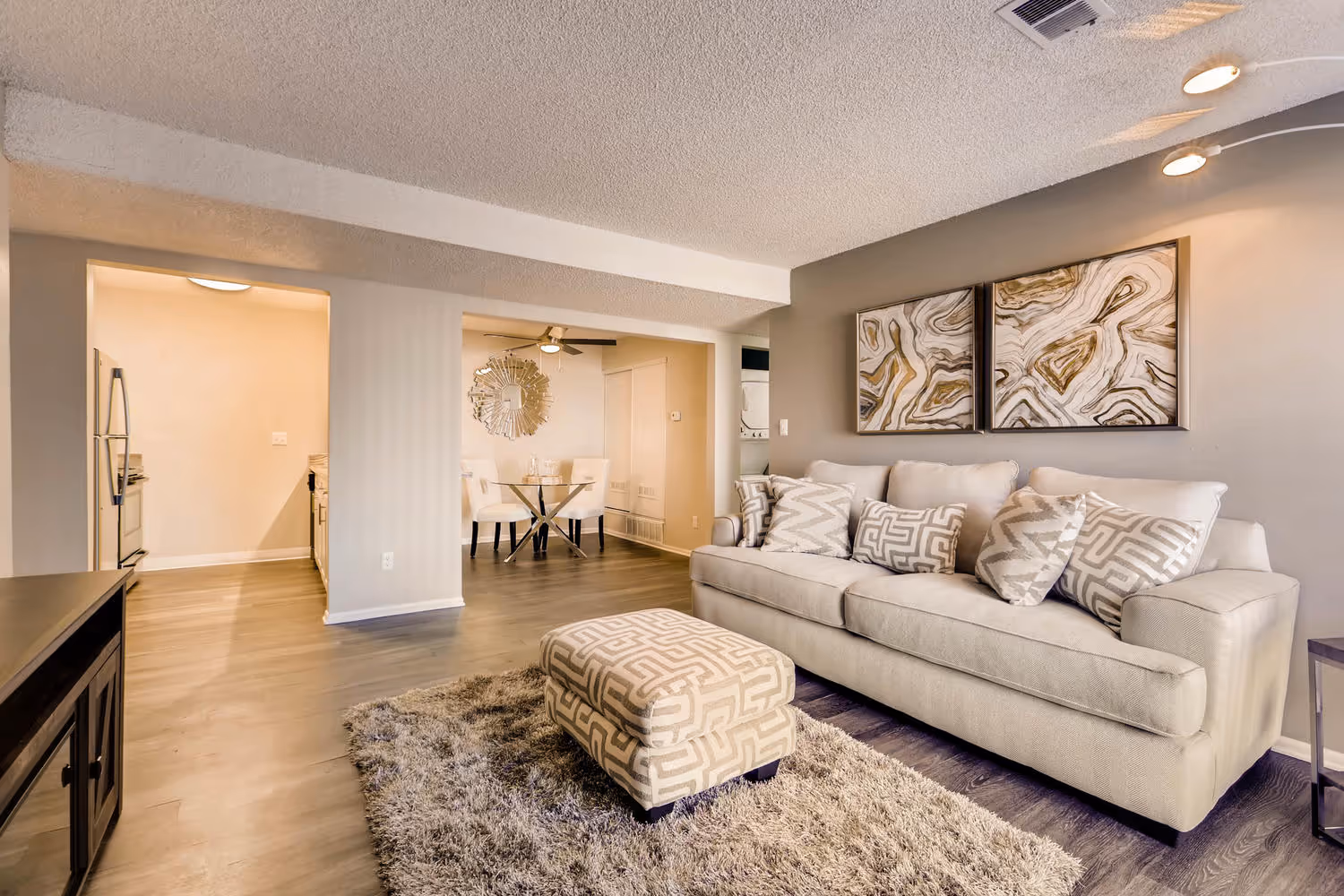 Open-concept living and dining area with a sofa, patterned ottoman, and hardwood-style floors, in an apartment at Villas at Holly in Centennial, CO.