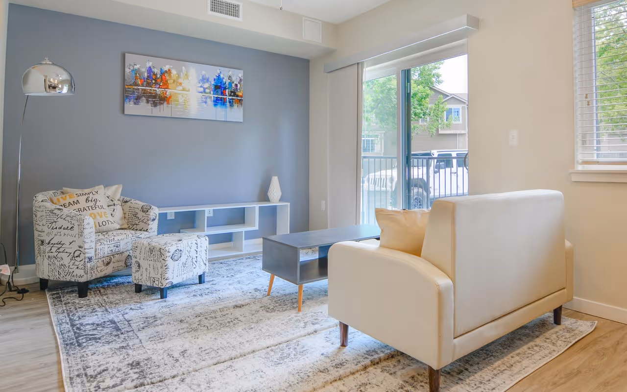 Bright apartment living room with a large sliding glass door, a sofa, and a patterned armchair, at Villas at Holly in Centennial, CO.