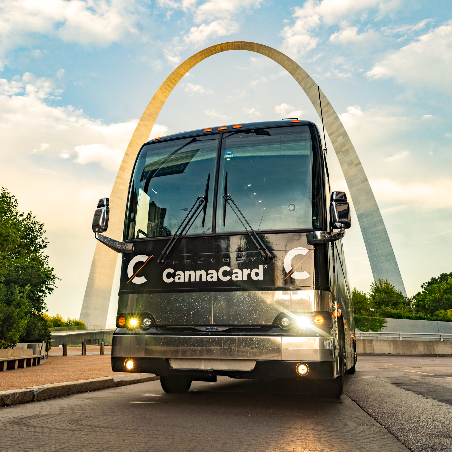 Front view of a black CannaCard bus parked with the Gateway Arch in the background under a partly cloudy sky.