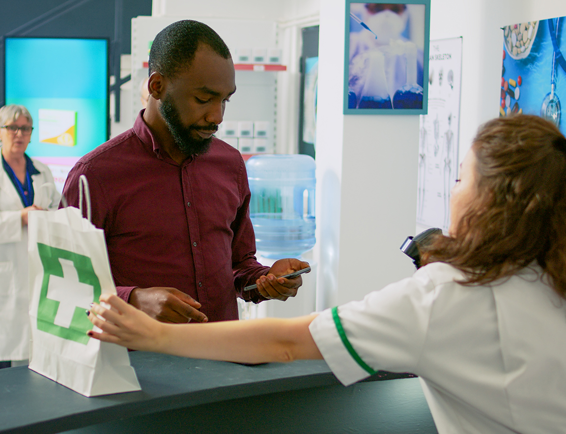 A man paying with his phone at a pharmacy counter while a pharmacist hands over a medicine bag with a green cross.