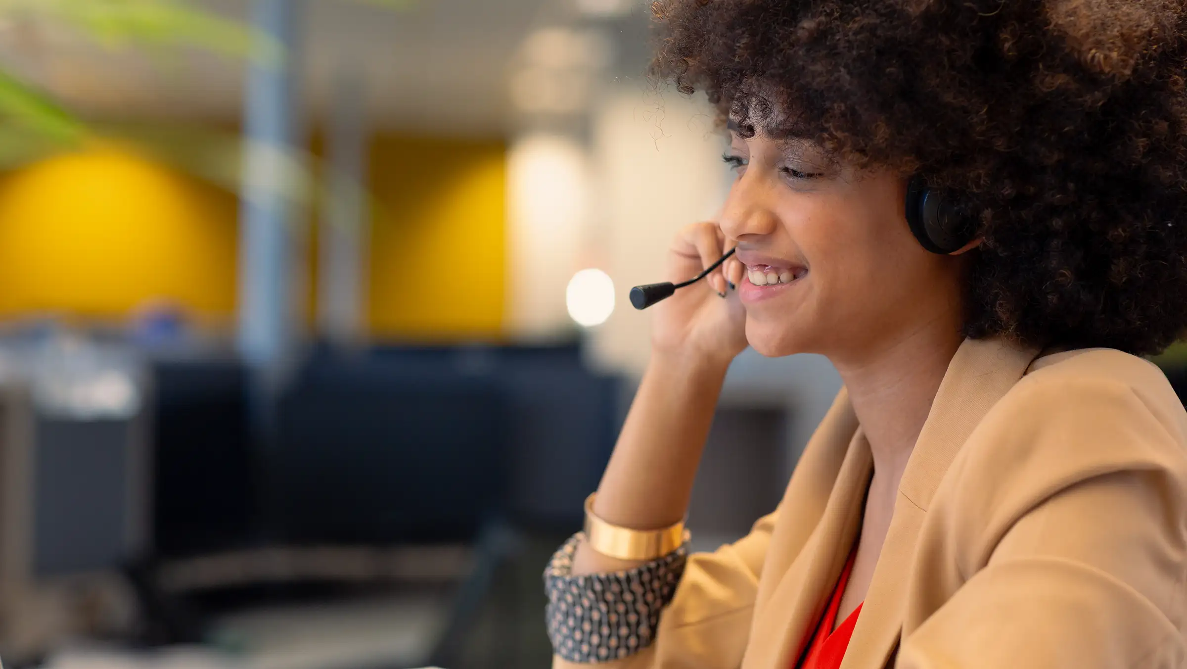 Smiling woman with curly hair wearing a headset and beige blazer, working in an office.
