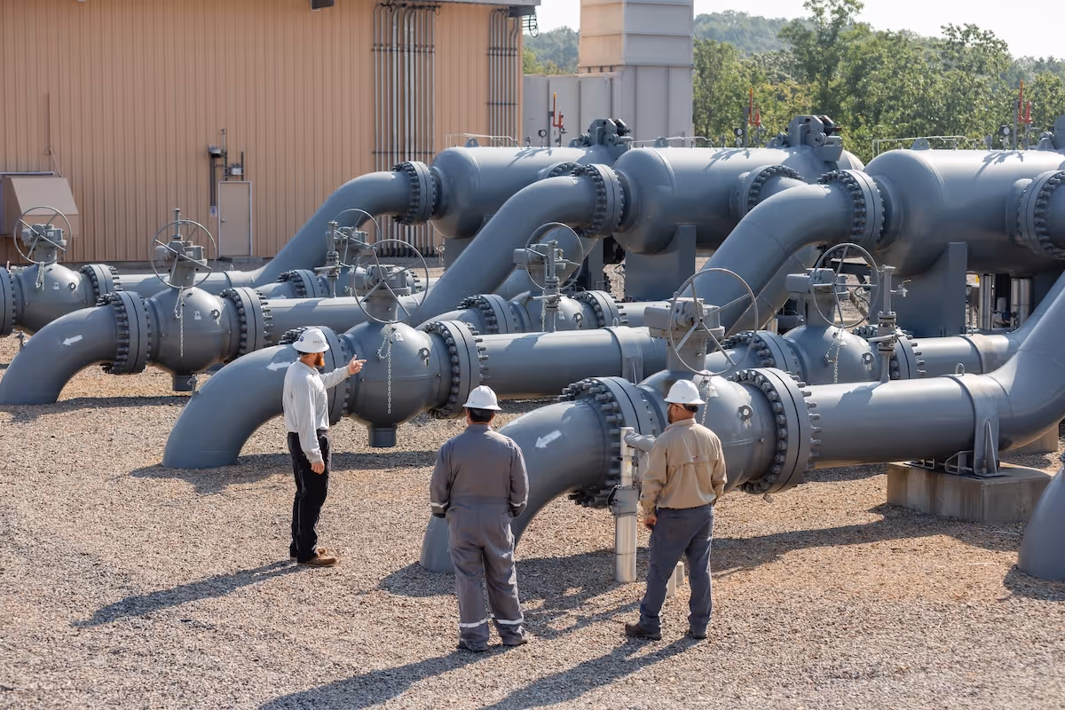Three workers in hard hats and safety gear stand on a gravel patch amidst a complex network of large gray pipelines and valves at an industrial facility
