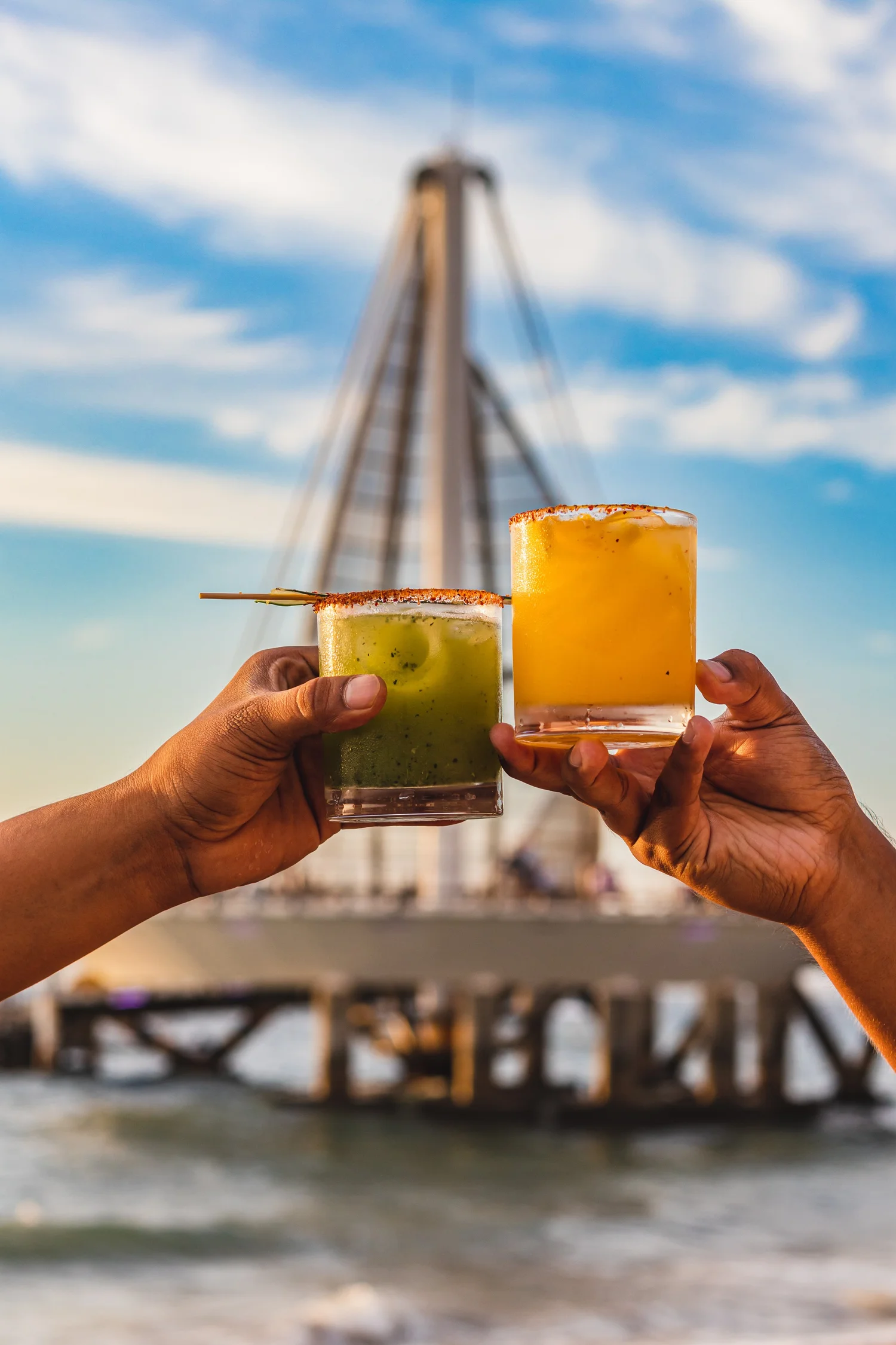Two cocktails raised on the beach in front of the Los Muertos Pier