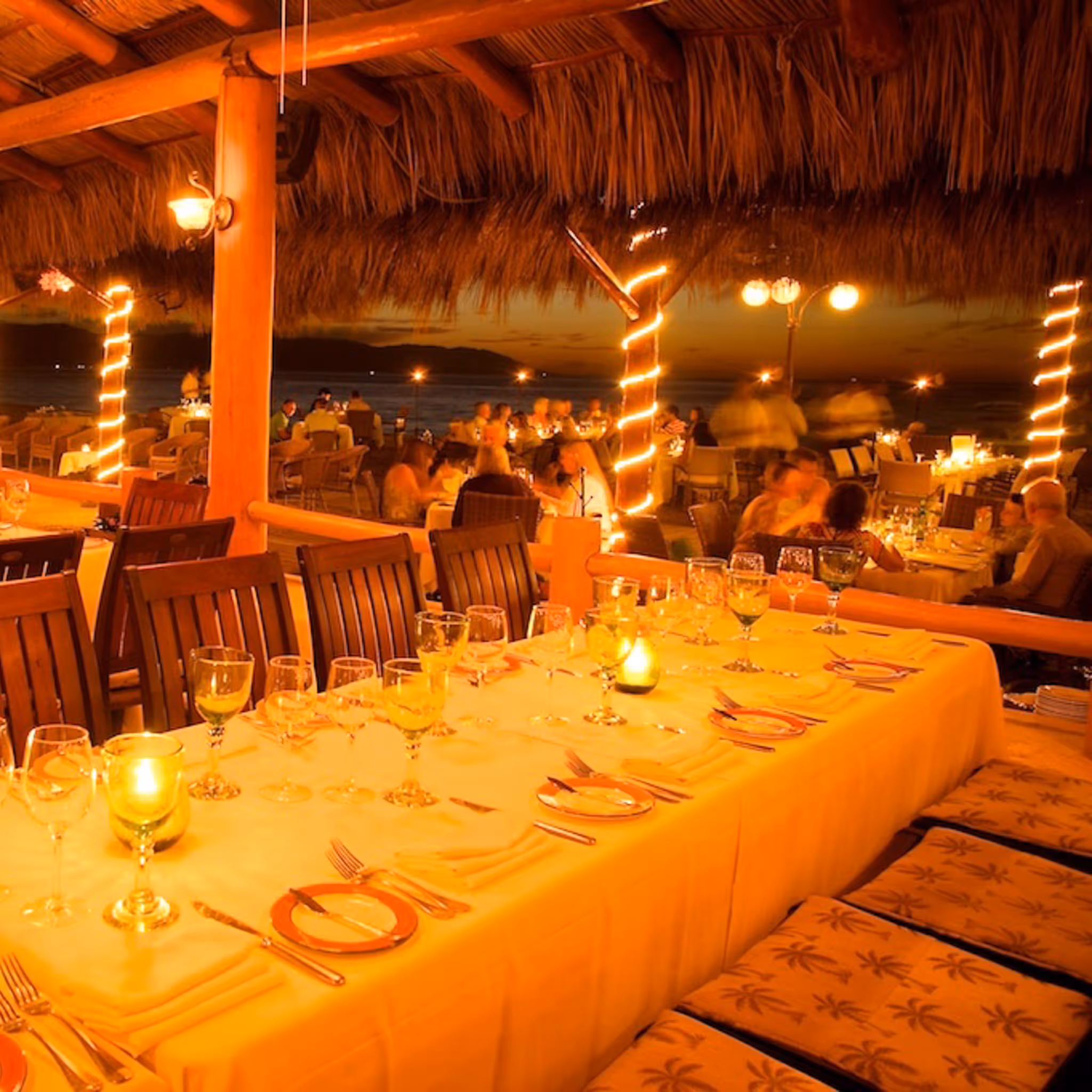 A long candlelit table set for a group dinner under the palapa roof