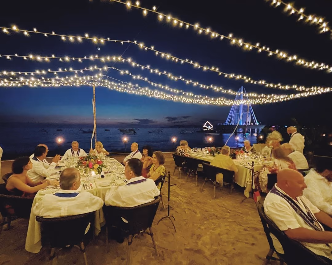 A hosted gala dinner at long tables on the sand under string lights, with the Los Muertos pier lit behind