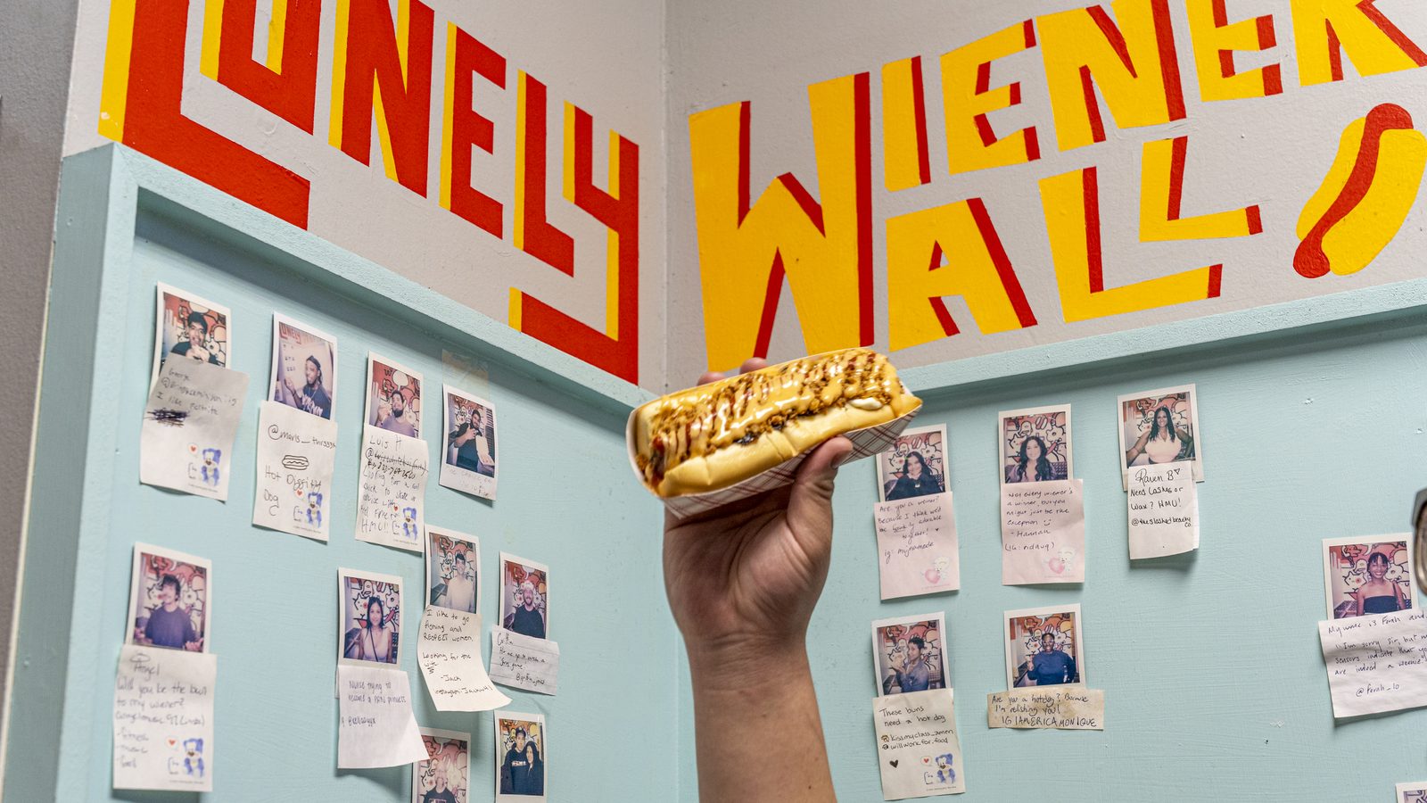 The Lonely Wiener Wall at Yoyo's Hotdog, hand-painted lettering above a wall of Polaroids, with a loaded hot dog held up in front of it