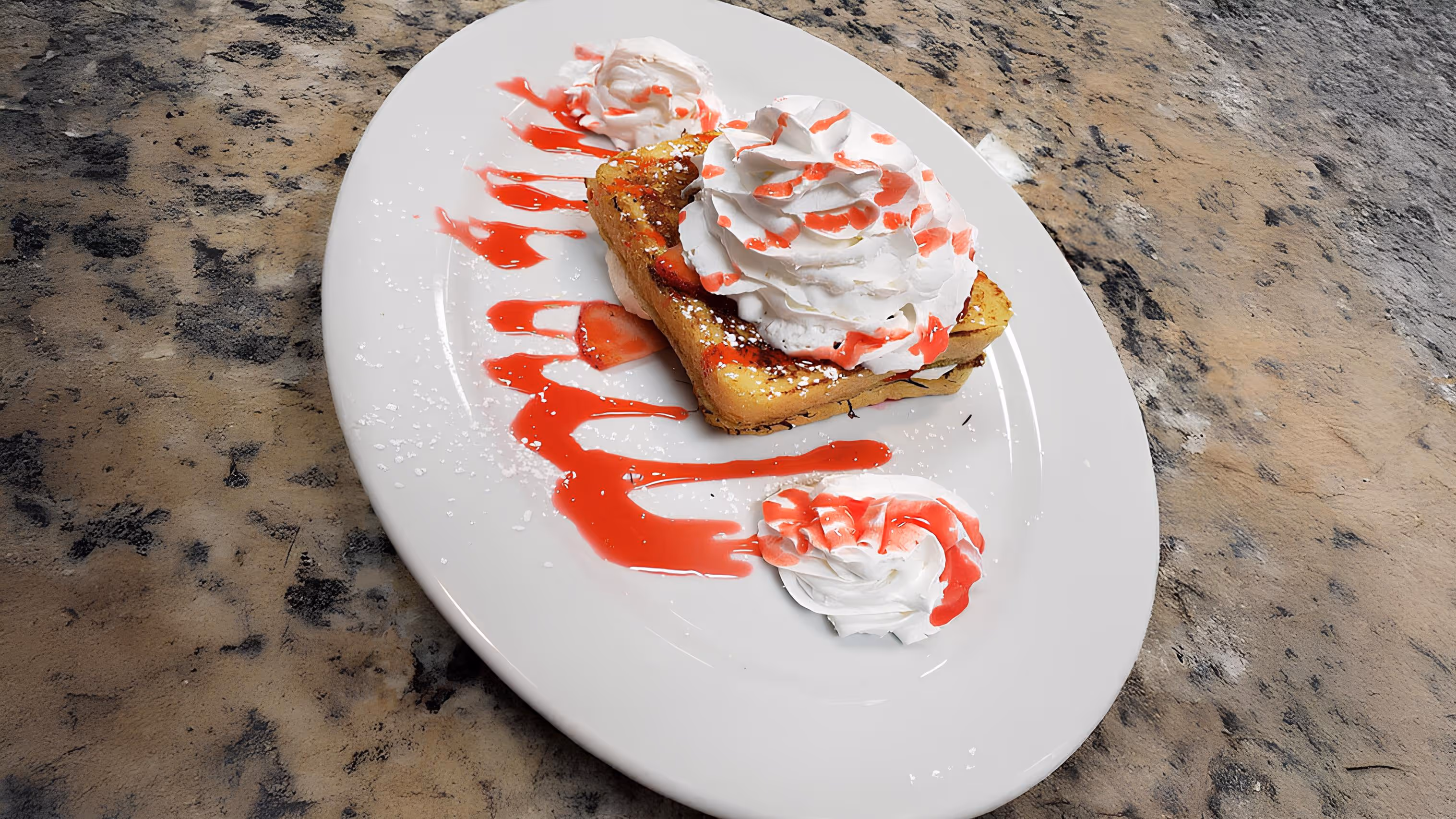 French toast topped with whipped cream, strawberries, and red syrup, on a white oval plate with dollops of cream.