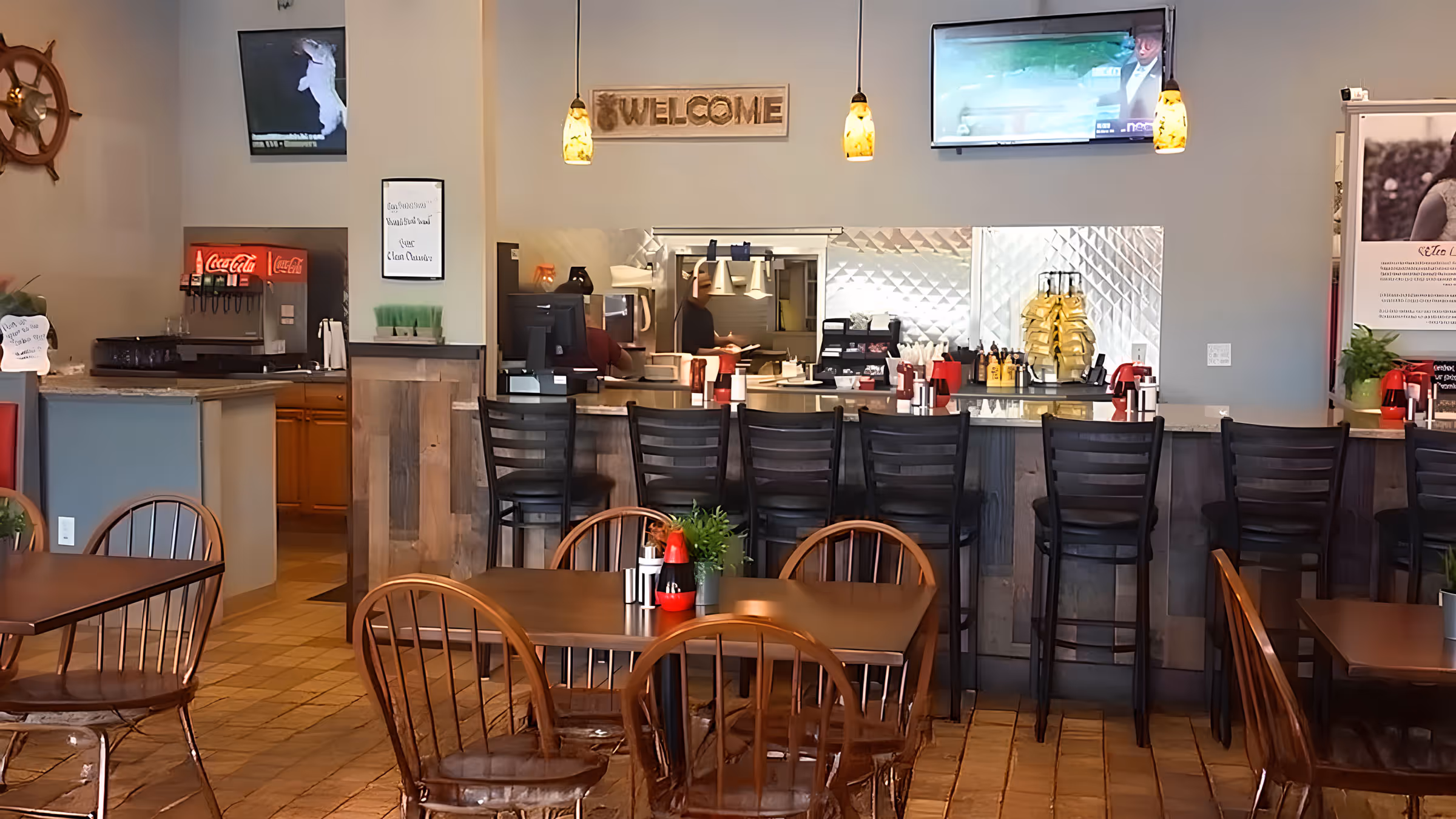 Cozy restaurant interior with wooden chairs and tables, a counter with high stools, a "Welcome" sign, and two TVs. Soft lighting creates a warm ambiance.