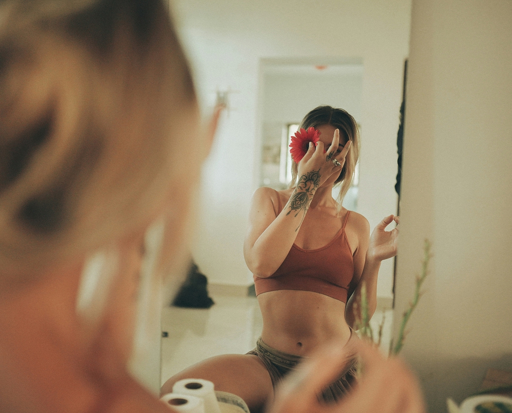 Woman wearing a rust-colored sports bra and green shorts sitting in front of a mirror holding a red flower in front of her face, revealing a hand tattoo.