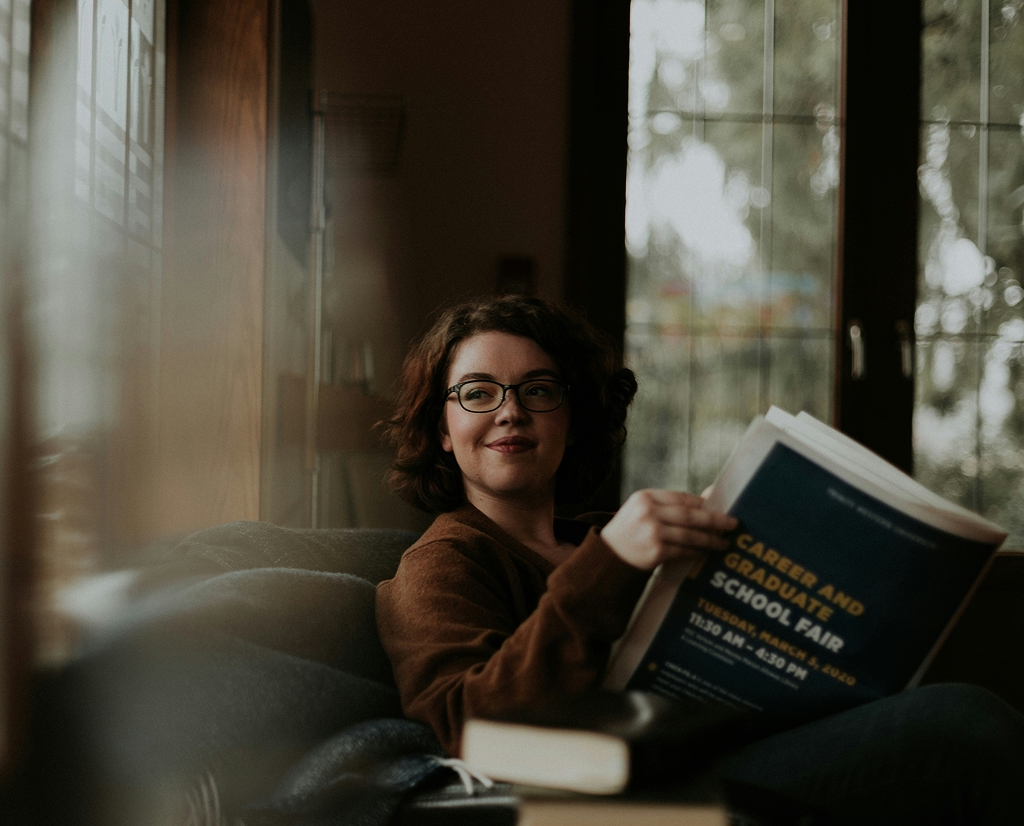 A woman with curly brown hair and glasses shares a relaxing moment indoors, smiling as she holds an open "Careers and Schools Fair" publication. The soft, natural light and nearby books create a cozy atmosphere, perfect for a mindful break with a cup of matcha.