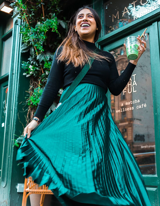 A woman with a joyous expression, wearing a black long-sleeved top and a vibrant, shimmering emerald green pleated skirt, stands outside a matcha wellness café. She holds a refreshing iced matcha latte, capturing a relaxing and mindful moment in the city.