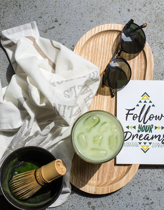A flat lay image of a relaxing matcha preparation scene on a grey textured surface. It includes a frothy matcha latte in a glass on a wooden tray, a traditional bamboo whisk in a black bowl, sunglasses, and a card that says "Follow Your Dreams."