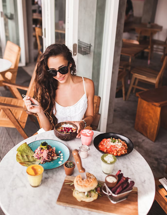 A woman with long curly hair and sunglasses smiles during a relaxing outdoor brunch. The marble table is filled with a colorful spread of healthy dishes and a vibrant green matcha latte, capturing a serene and mindful dining moment.
