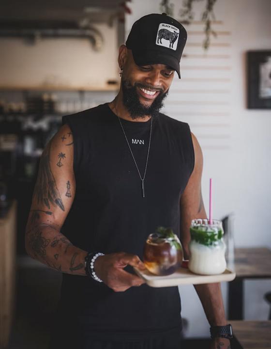 A smiling man in a black sleeveless shirt and a baseball cap stands in a cafe, holding a wooden tray with two drinks. One is a dark iced coffee, and the other is a vibrant layered matcha latte, symbolizing a relaxing break focused on health and weight loss.